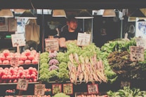 A market stall brimming with fresh produce such as tomatoes, carrots, cauliflower, and various leafy greens. Handwritten price signs are visible throughout the display. A vendor stands behind the counter, partially obscured by the vegetables.