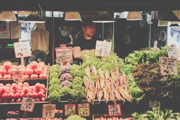 A market stall brimming with fresh produce such as tomatoes, carrots, cauliflower, and various leafy greens. Handwritten price signs are visible throughout the display. A vendor stands behind the counter, partially obscured by the vegetables.