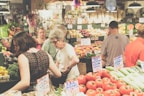 A bustling indoor farmers market features several people examining fresh produce and interacting with vendors. The produce is attractively displayed with signs indicating prices and varieties, and includes a variety of vegetables like tomatoes, onions, and avocados. The environment is lively and colorful, with an abundance of fresh vegetables and fruits arranged neatly on the stalls.