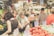 A bustling indoor farmers market features several people examining fresh produce and interacting with vendors. The produce is attractively displayed with signs indicating prices and varieties, and includes a variety of vegetables like tomatoes, onions, and avocados. The environment is lively and colorful, with an abundance of fresh vegetables and fruits arranged neatly on the stalls.