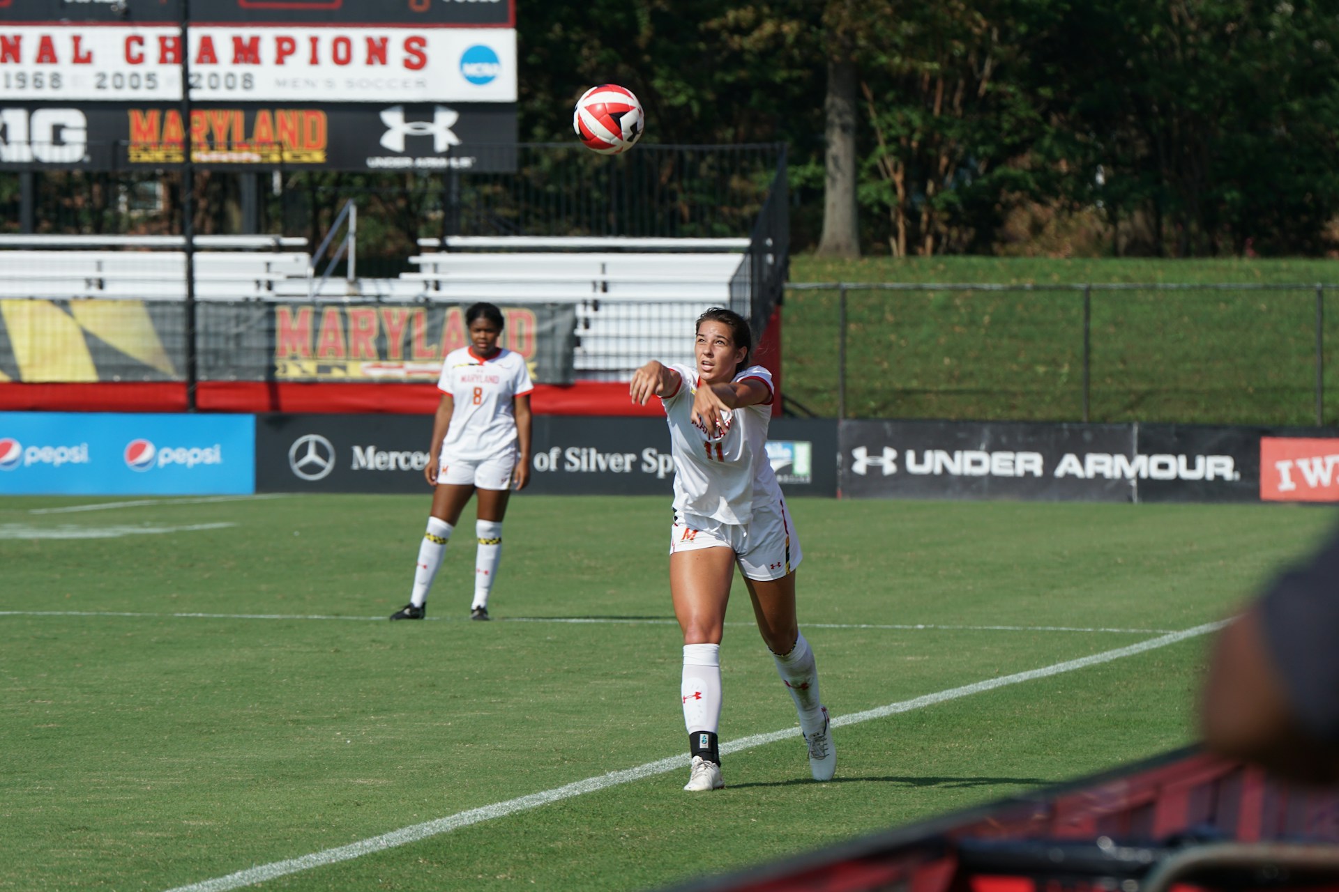 A female soccer player in white uniform is throwing a soccer ball during a match. Another player in a white uniform is standing in the background. The scene is set on a green soccer field with advertisements and banners, including 'Maryland' and 'National Champions', visible in the background.
