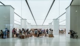 A modern, brightly lit store interior with numerous people sitting on wooden stools, possibly in a waiting area or workshop. Customers appear to be engaged with electronic devices. The setting has a minimalist design with white walls and floors, and an overall sleek appearance.