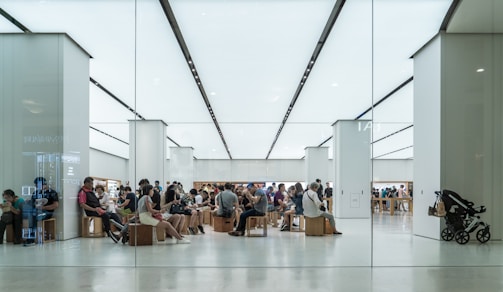 A modern, brightly lit store interior with numerous people sitting on wooden stools, possibly in a waiting area or workshop. Customers appear to be engaged with electronic devices. The setting has a minimalist design with white walls and floors, and an overall sleek appearance.
