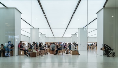 A modern, brightly lit store interior with numerous people sitting on wooden stools, possibly in a waiting area or workshop. Customers appear to be engaged with electronic devices. The setting has a minimalist design with white walls and floors, and an overall sleek appearance.