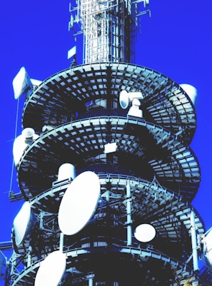 A large communication tower with multiple satellite dishes and antennas is set against a clear blue sky. The structure is composed of metal girders and platforms, with various white satellite dishes mounted at different levels.