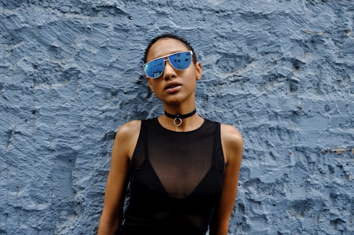 A person wearing reflective sunglasses and a black sleeveless top stands against a textured blue wall. The sunglasses reflect a cityscape, adding an urban vibe. A choker necklace with a ring accentuates the outfit, contributing to the edgy and stylish appearance.