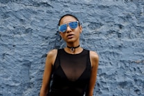 A person wearing reflective sunglasses and a black sleeveless top stands against a textured blue wall. The sunglasses reflect a cityscape, adding an urban vibe. A choker necklace with a ring accentuates the outfit, contributing to the edgy and stylish appearance.