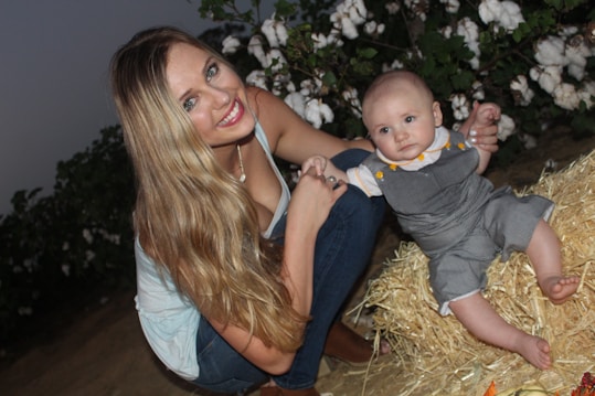 A woman with long blonde hair, wearing a light blue top and jeans, smiles warmly while crouching beside a baby. The baby is sitting on a pile of straw, dressed in a gray outfit with a white collar and yellow buttons. Cotton plants with white flowers are in the background, suggesting an outdoor setting.