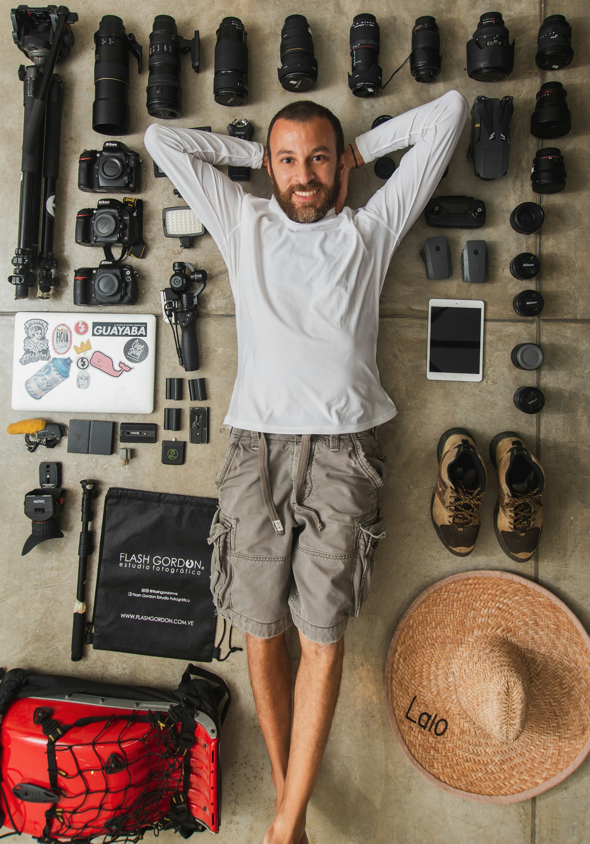 A man is lying on the floor surrounded by an organized collection of photography gear, including multiple camera lenses, camera bodies, a tripod, and various accessories. He has a relaxed demeanor, wearing a white long-sleeved shirt and cargo shorts. Nearby, there are hiking shoes and a large straw hat with the word 'Lalo' written on it.