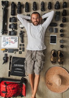 A man is lying on the floor surrounded by an organized collection of photography gear, including multiple camera lenses, camera bodies, a tripod, and various accessories. He has a relaxed demeanor, wearing a white long-sleeved shirt and cargo shorts. Nearby, there are hiking shoes and a large straw hat with the word 'Lalo' written on it.