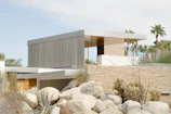 A modern architectural structure featuring flat roofs and clean, geometric lines is surrounded by natural desert landscaping. The building incorporates natural stone and metal materials, blending harmoniously with the arid environment. Agave plants and boulders are prominent in the foreground, and palm trees are visible in the background, emphasizing the desert setting.