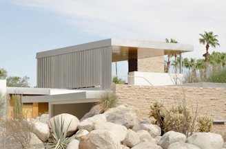 A modern architectural structure featuring flat roofs and clean, geometric lines is surrounded by natural desert landscaping. The building incorporates natural stone and metal materials, blending harmoniously with the arid environment. Agave plants and boulders are prominent in the foreground, and palm trees are visible in the background, emphasizing the desert setting.