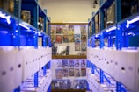 Rows of shelves filled with white storage boxes line the sides, with colorful comic book covers displayed on the wall at the far end. The shelving units are painted bright blue, contrasting with the neutral tones of the boxes and the vivid colors of the comics.