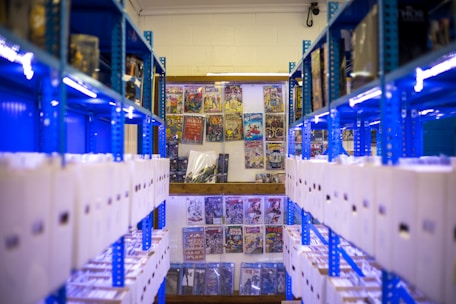 Rows of shelves filled with white storage boxes line the sides, with colorful comic book covers displayed on the wall at the far end. The shelving units are painted bright blue, contrasting with the neutral tones of the boxes and the vivid colors of the comics.