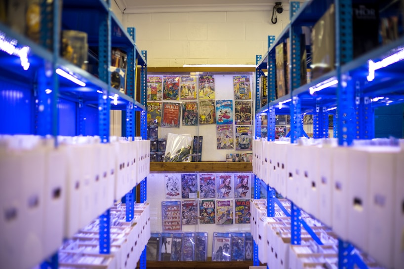 Rows of shelves filled with white storage boxes line the sides, with colorful comic book covers displayed on the wall at the far end. The shelving units are painted bright blue, contrasting with the neutral tones of the boxes and the vivid colors of the comics.