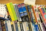 A collection of audio cassette tapes and books are arranged on a shelf. These include titles from various genres, such as mysteries, self-help, and classic literature. The tapes are stacked at different angles, creating a colorful and eclectic display with visible titles like 'Great Expectations,' 'Gulliver's Travels,' and 'The Cat Who Brought Down the House.'