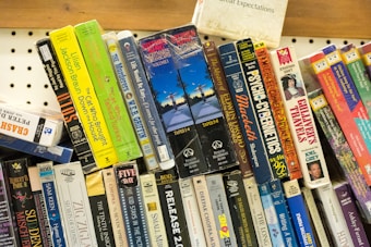 A collection of audio cassette tapes and books are arranged on a shelf. These include titles from various genres, such as mysteries, self-help, and classic literature. The tapes are stacked at different angles, creating a colorful and eclectic display with visible titles like 'Great Expectations,' 'Gulliver's Travels,' and 'The Cat Who Brought Down the House.'