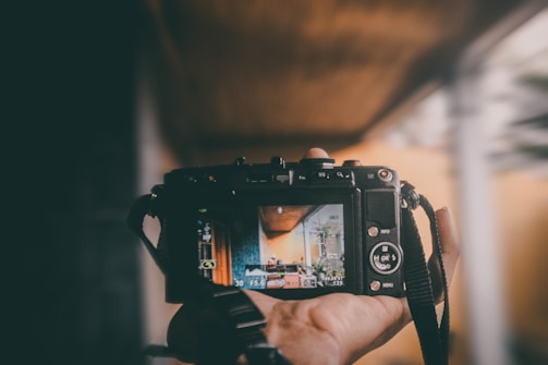 A hand holds a digital camera, capturing a view of an interior room with plants and warm lighting. The camera's screen displays the room being photographed, showing elements such as a couch and decorations.