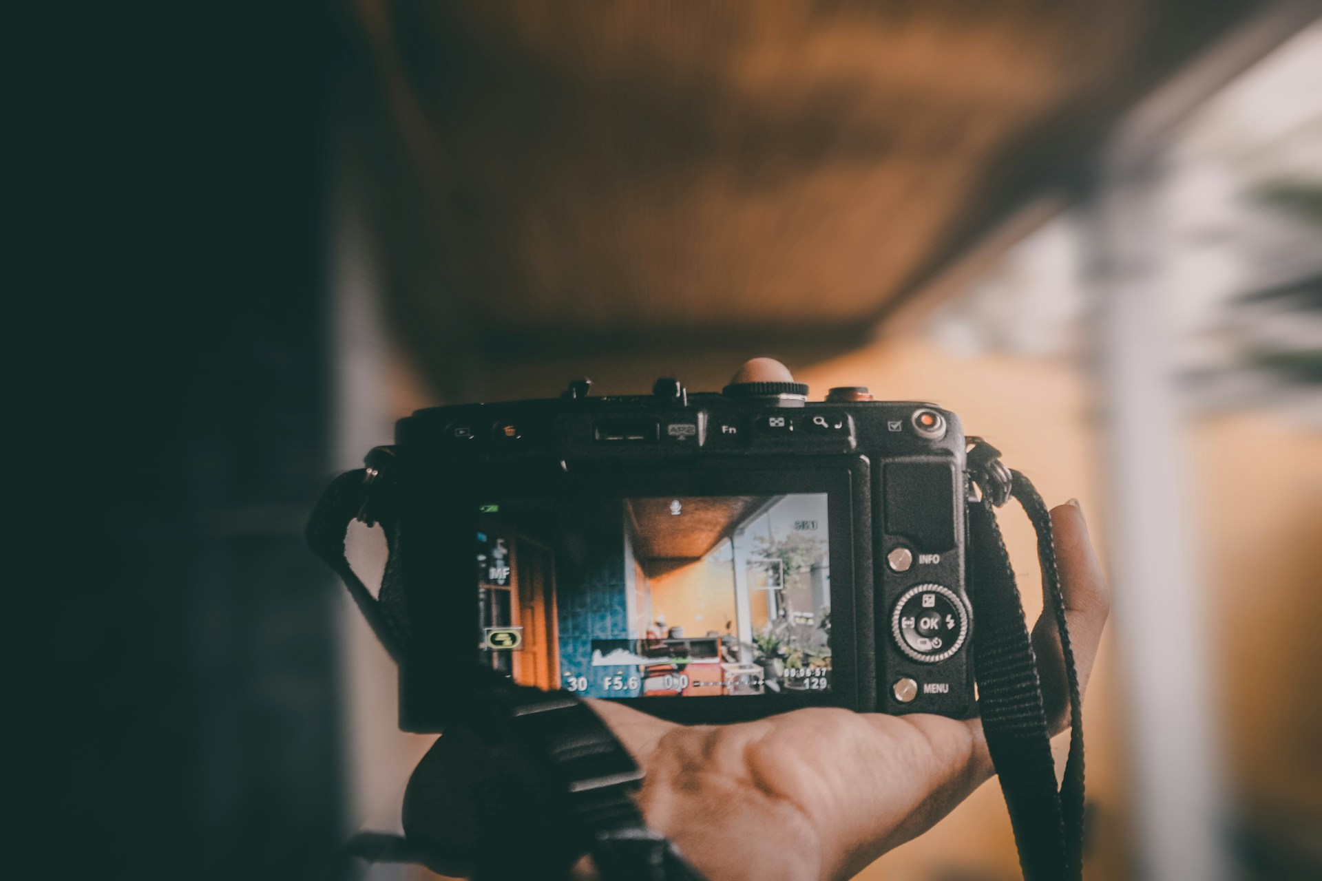 A hand holds a digital camera, capturing a view of an interior room with plants and warm lighting. The camera's screen displays the room being photographed, showing elements such as a couch and decorations.