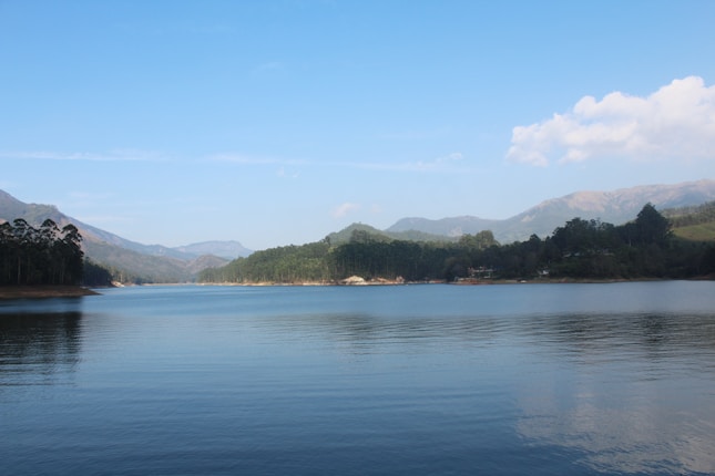 A serene lake with calm water reflecting the surrounding landscape of forested hills and distant mountains under a clear blue sky.