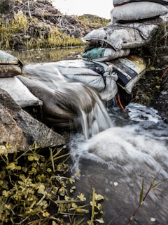 A stream of water flows over a makeshift dam constructed from stacked sandbags. The water cascades down smoothly, with vegetation and small plants growing around the water's edge. The scene is set outdoors in a natural environment with earthy tones.