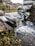 A stream of water flows over a makeshift dam constructed from stacked sandbags. The water cascades down smoothly, with vegetation and small plants growing around the water's edge. The scene is set outdoors in a natural environment with earthy tones.