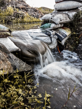 A stream of water flows over a makeshift dam constructed from stacked sandbags. The water cascades down smoothly, with vegetation and small plants growing around the water's edge. The scene is set outdoors in a natural environment with earthy tones.