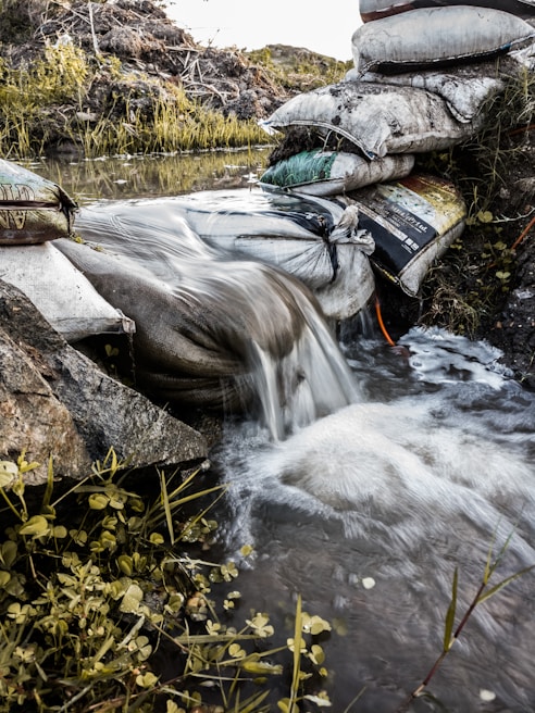 A stream of water flows over a makeshift dam constructed from stacked sandbags. The water cascades down smoothly, with vegetation and small plants growing around the water's edge. The scene is set outdoors in a natural environment with earthy tones.