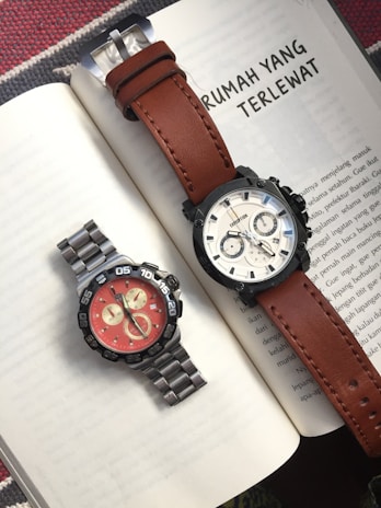 Two wristwatches are placed on an open book, one with a brown leather strap and a light dial, and the other with a metallic strap and a bright red dial. The background features text in the Indonesian language.