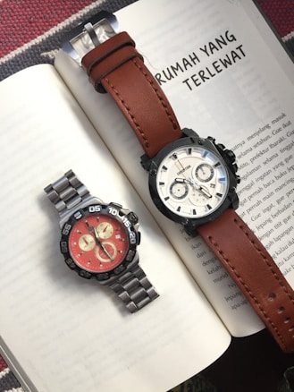 Two wristwatches are placed on an open book, one with a brown leather strap and a light dial, and the other with a metallic strap and a bright red dial. The background features text in the Indonesian language.