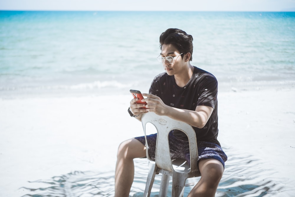 man in black shirt and blue board shorts sitting on plastic chair near seashore