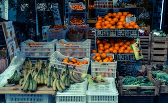 A market stall displaying a variety of fresh fruits and vegetables. Crates filled with bright oranges, ripe bananas, and assorted greens are arranged neatly. Signage with Greek writing is visible, and there are garlic bulbs hanging in the background.