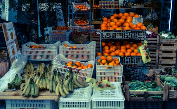 A market stall displaying a variety of fresh fruits and vegetables. Crates filled with bright oranges, ripe bananas, and assorted greens are arranged neatly. Signage with Greek writing is visible, and there are garlic bulbs hanging in the background.
