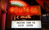 Neon-lit signage featuring 'Route 66' in bright orange and yellow lights above a marquee with the words 'Creature from the Black Lagoon.' An illuminated arrow labeled 'Drive-In' points downward.