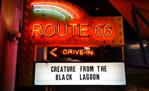 Neon-lit signage featuring 'Route 66' in bright orange and yellow lights above a marquee with the words 'Creature from the Black Lagoon.' An illuminated arrow labeled 'Drive-In' points downward.