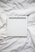 A white book with the word 'minimalismus' printed in bold black letters on the cover, placed on a white, crumpled cloth or bedspread.