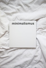 A white book with the word 'minimalismus' printed in bold black letters on the cover, placed on a white, crumpled cloth or bedspread.