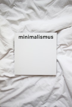 A white book with the word 'minimalismus' printed in bold black letters on the cover, placed on a white, crumpled cloth or bedspread.