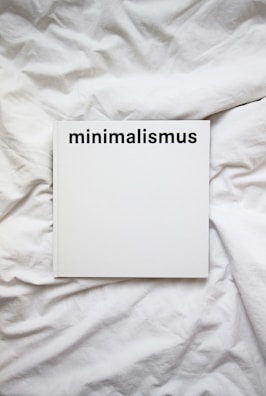 A white book with the word 'minimalismus' printed in bold black letters on the cover, placed on a white, crumpled cloth or bedspread.