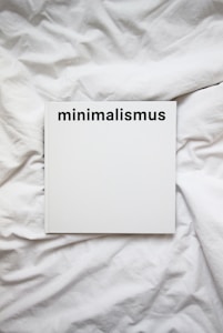 A white book with the word 'minimalismus' printed in bold black letters on the cover, placed on a white, crumpled cloth or bedspread.