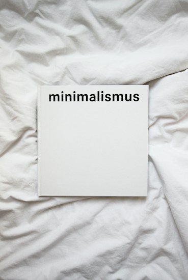 A white book with the word 'minimalismus' printed in bold black letters on the cover, placed on a white, crumpled cloth or bedspread.