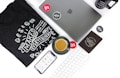 An arrangement of technology and design-related items on a white surface. Includes a laptop with the Apple logo, a smartphone displaying a design app interface, a black t-shirt featuring the phrase 'Design Makes Everything Possible', a mug with a similar design-themed phrase, a keyboard, and a saucer with a coffee cup. Various stickers and a black notebook are also present.
