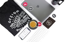 An arrangement of technology and design-related items on a white surface. Includes a laptop with the Apple logo, a smartphone displaying a design app interface, a black t-shirt featuring the phrase 'Design Makes Everything Possible', a mug with a similar design-themed phrase, a keyboard, and a saucer with a coffee cup. Various stickers and a black notebook are also present.