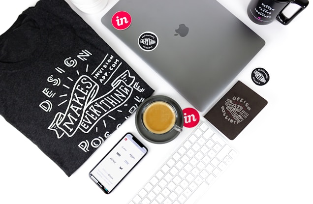 An arrangement of technology and design-related items on a white surface. Includes a laptop with the Apple logo, a smartphone displaying a design app interface, a black t-shirt featuring the phrase 'Design Makes Everything Possible', a mug with a similar design-themed phrase, a keyboard, and a saucer with a coffee cup. Various stickers and a black notebook are also present.