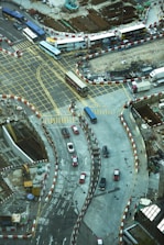A busy urban intersection with multiple lanes marked by yellow grid lines and traffic movement. Buses and cars are navigating the area, surrounded by construction zones with red and white barriers. The construction site features visible dirt and machinery, while roads are marked with directional arrows and bilingual text.