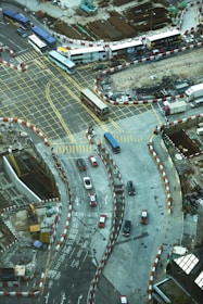 A busy urban intersection with multiple lanes marked by yellow grid lines and traffic movement. Buses and cars are navigating the area, surrounded by construction zones with red and white barriers. The construction site features visible dirt and machinery, while roads are marked with directional arrows and bilingual text.