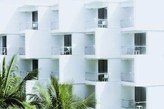 White modern building with glass balconies featuring outdoor furniture, and lush green palm fronds in the foreground.