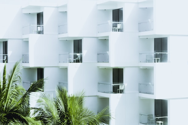 White modern building with glass balconies featuring outdoor furniture, and lush green palm fronds in the foreground.