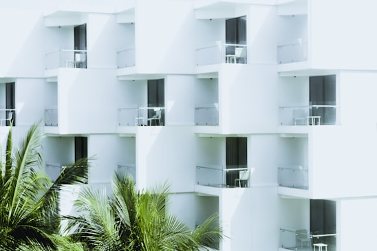 White modern building with glass balconies featuring outdoor furniture, and lush green palm fronds in the foreground.