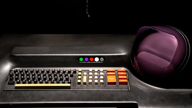 A retro-style keyboard and control panel with a set of colored buttons, a series of white, yellow, and orange buttons labeled 'SEND,' 'PRINT,' and others. To the right, there is a rounded, purple screen or dome on a dark background.